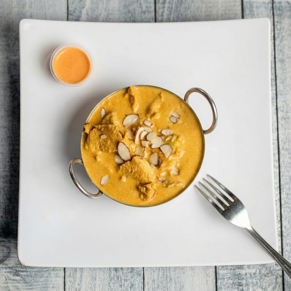 Chicken curry with sliced almonds in a metal bowl, with a side of sauce and a fork.