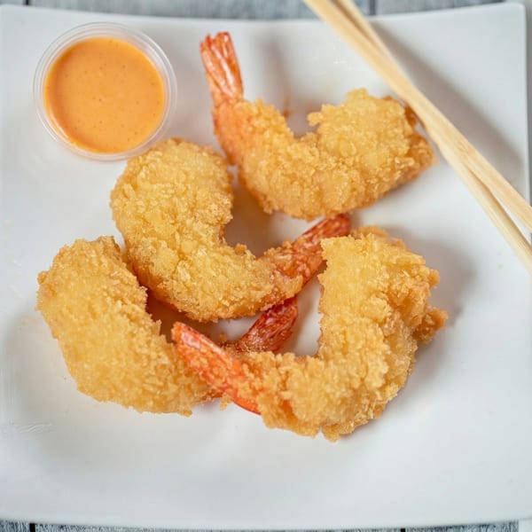 Four golden-brown fried shrimp with tails on, a side of orange dipping sauce, and chopsticks.