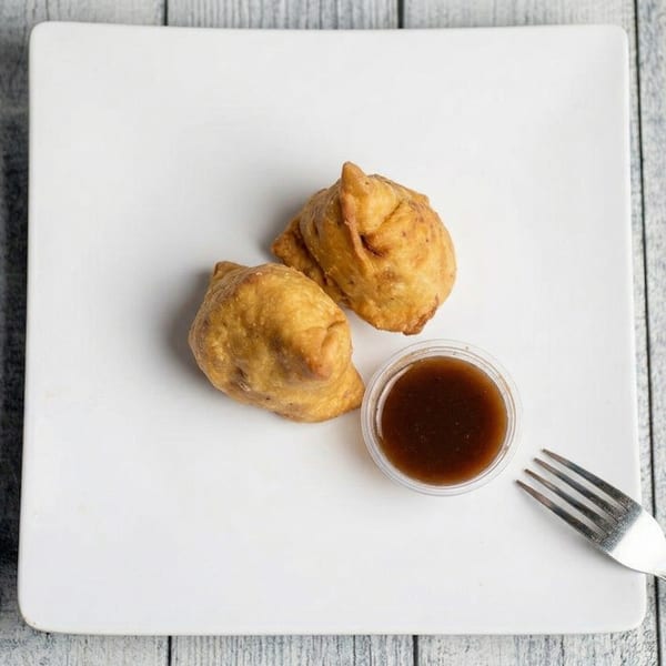 Two golden-brown samosas with a small cup of dipping sauce and a fork on a white plate.