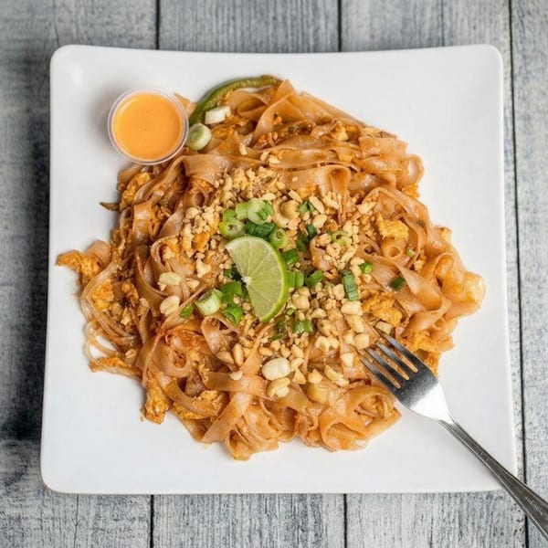 Pad Thai noodles with peanuts, green onions, lime, and a side of sauce on a white plate.