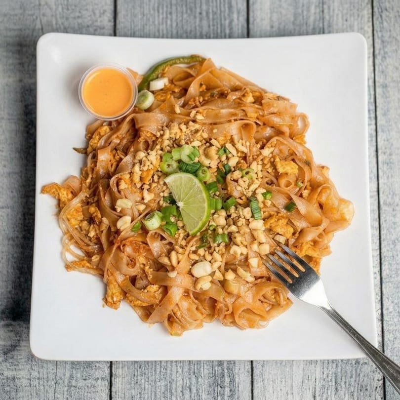 Pad Thai noodles with peanuts, green onions, lime, and a side of sauce on a white plate.